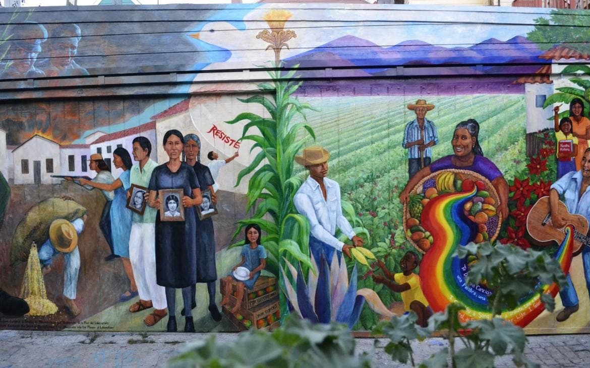 Community mural depicting Latin American life: women holding framed portraits, farmworkers in a green field, a woman carrying a basket of fruit spilling a rainbow, and a guitarist